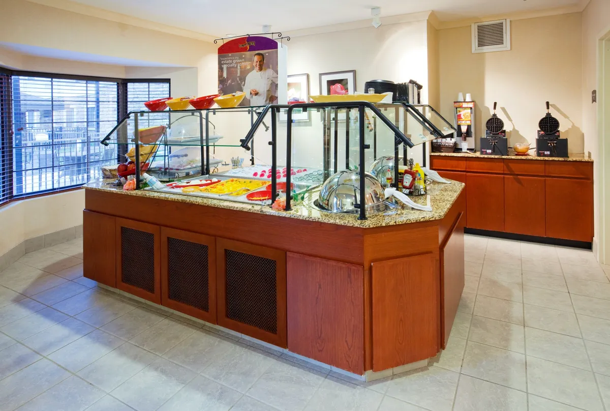 This image shows a buffet counter with a glass display, various food items, a waffle maker, and decorative artwork above. The scene is set up for guests to socialize over breakfast or casual meals, with an inviting and well-lit atmosphere.