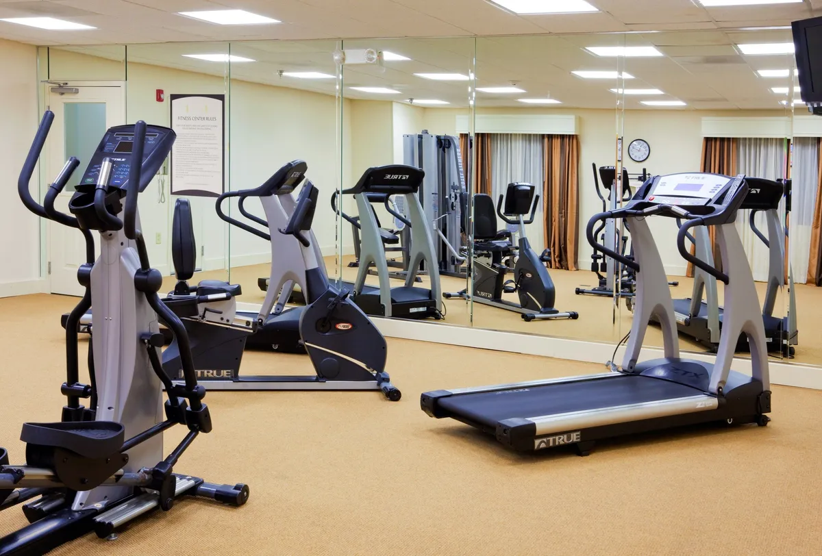 A bright, modern fitness center featuring an elliptical machine, treadmill, and weight machine, all set against a mirrored wall. The space is designed for both individual workouts and group fitness classes, creating an energetic and inviting environment for staying active.