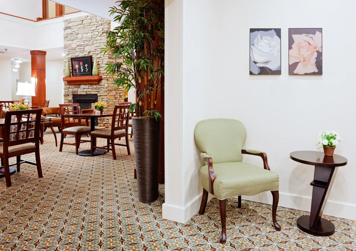 A cozy lounge area featuring a stylish green chair and small round table set beside a modern fireplace. Artwork adds an element of sophistication to the walls, and a leafy potted plant brings a touch of nature indoors. The space conveys a comfortable atmosphere ideal for reading, studying, or enjoying a quiet moment.