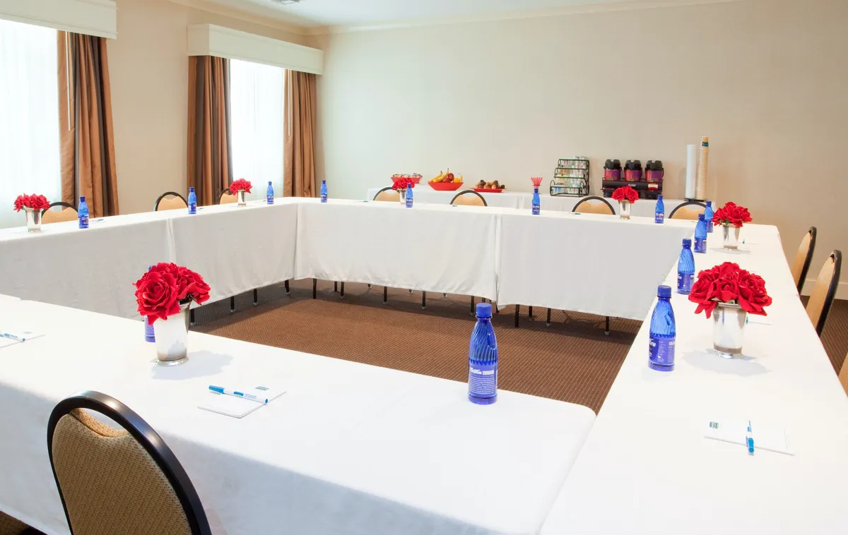 This interior shot features a rectangular dining table arrangement adorned with fresh red roses and blue water bottles, set up for group gatherings or meals. The table is situated in a bright, organized space suited for both work and dining.