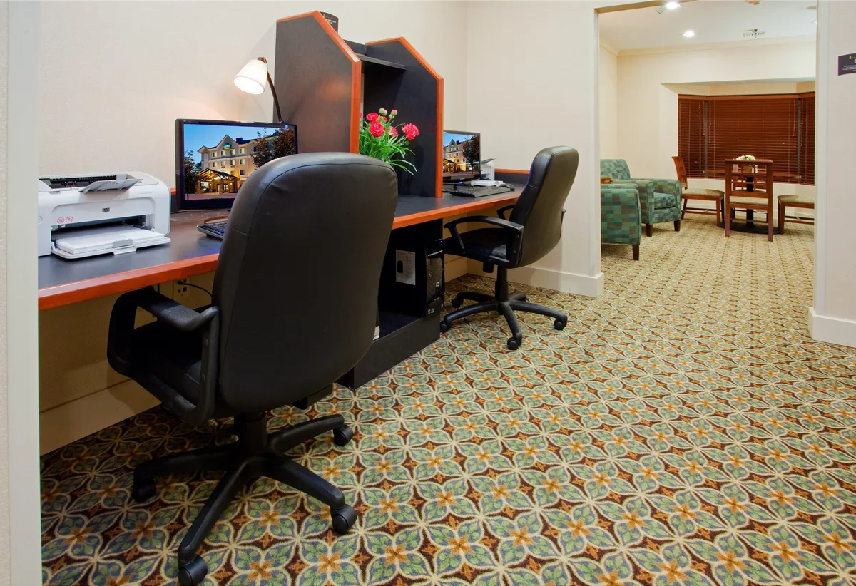 This image presents a functional workspace area featuring two computer desks, a printer, a floral arrangement, and a comfortable seating area in the background. The overall scene is bright and organized, suggesting a productive yet welcoming environment ideal for work or study.