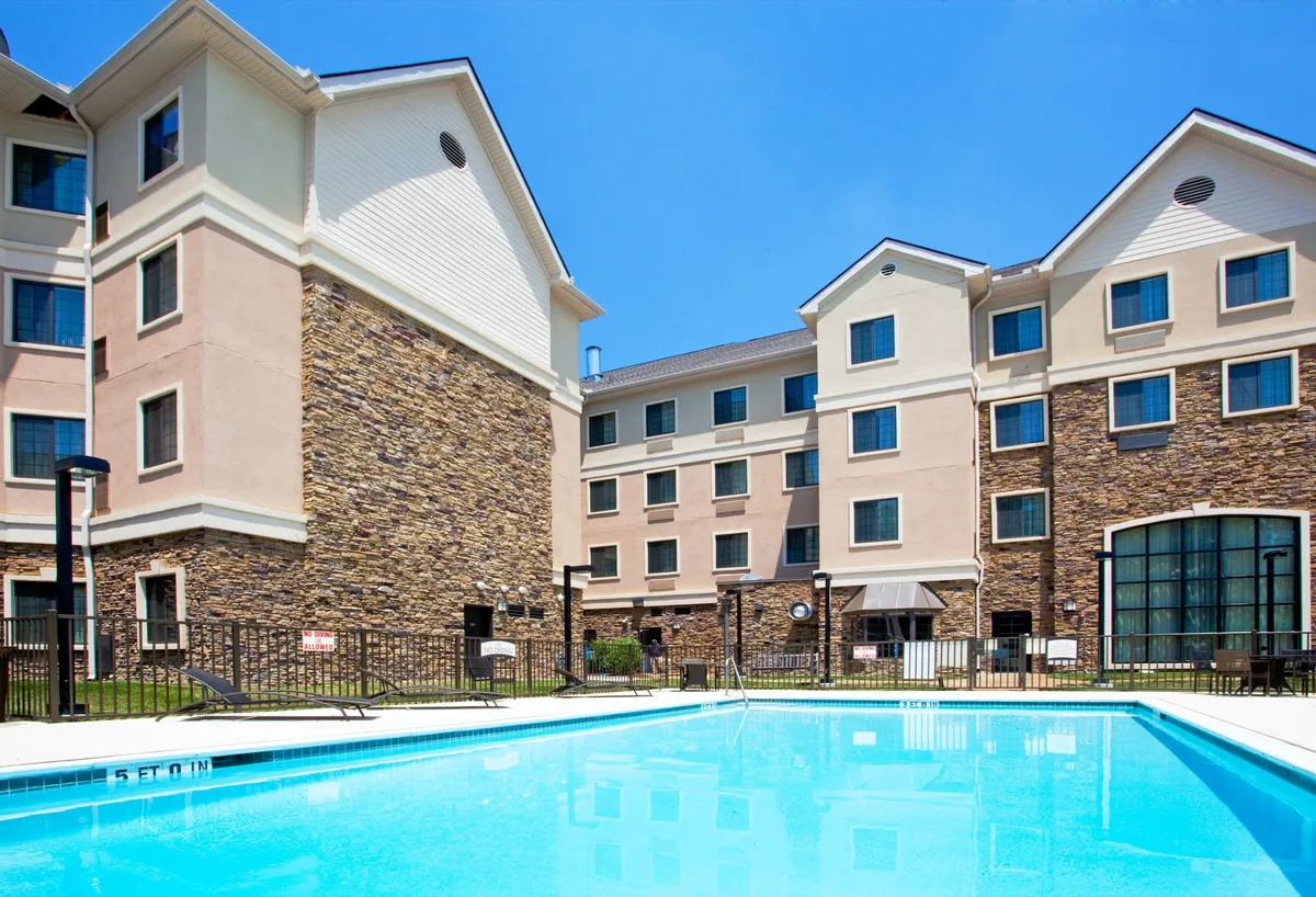An inviting outdoor swimming pool surrounded by a landscaped area and a stone-facade hotel building. The scene includes sunbathers and swimmers, capturing the pool's appeal during sunset and highlighting its relaxing atmosphere.