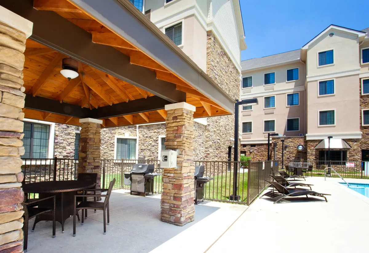 A vibrant outdoor area combining a BBQ grill, comfortable seating, and a view of the swimming pool, all enclosed by lush greenery. The space is set up for social gatherings, barbecuing, and relaxing afternoons by the pool.