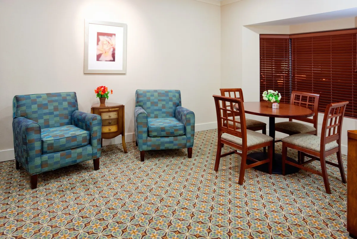 The interior dining/living area features blue patterned chairs, a round dining table topped with a floral centerpiece, and tasteful framed artwork. The mood is comfortable and inviting, with natural light and a sense of space for both relaxation and gathering.