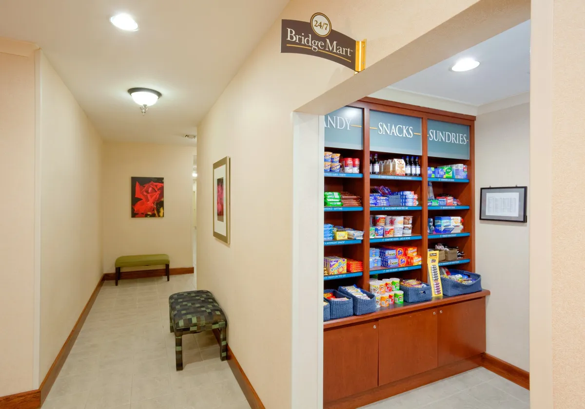 A bright, accessible convenience area with the 'Bridge Mart' sign above a well-stocked snack display, complemented by comfortable benches and vibrant artwork. The space is designed for quick stops and easy access to refreshments, supporting guests’ busy schedules.